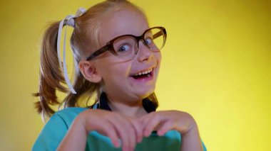 Gözlük takan, ağzı açık ve elleri göğsüne yakın bir şekilde heyecanlı bir sürpriz yapan bir çocuğun yakın çekimi. Sarı örgülü ve büyük kahverengi gözlüklü neşeli, genç bir kız öğrenci canlı sarı bir arka plana yürekten gülüyor..