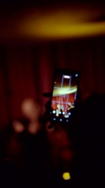 Dikey. Bir anı yakalamak: Smartphone, zengin kırmızı perdelere karşı loş, samimi bir canlı müzik performansı kaydediyor. Siluetli seyirci elinde telefonla sahnede müzisyenlerin görüntüsüyle ekranda parlıyordu..