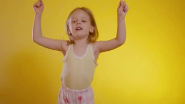Uzun sarı saçlı neşeli genç kız canlı, güneşli bir arka planda dans ediyor, açık sarı bir bluz ve desenli şort giyiyor. Çocuk heyecanlı bir anda iki yumruğunu birden kaldırıyor. 