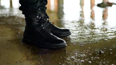Bulanık bir su birikintisinin kenarında duran siyah çizmeler nemli ve kentsel ortamda yukarıdaki ışığı yansıtıyor. Dalgalanan suyun yanında koyu renk pantolonlara ve dantelli botlara yakın çekim. Yağmur damlaları küçük lekeler oluşturur..