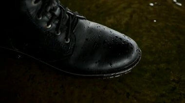 Su dalgalarının yanında sağlam, bağcıklı siyah botlar. Siyah deri botlar sığ ve yansıtıcı bir su birikintisinin kenarında duruyor. Bulanık ışıkların altında ıslak betonla buluşan eskimiş botlar..