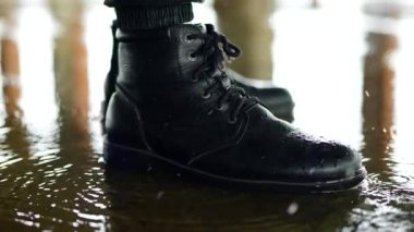 Su dalgalarının yanında sağlam, bağcıklı siyah botlar. Siyah deri botlar sığ ve yansıtıcı bir su birikintisinin kenarında duruyor. Bulanık ışıkların altında ıslak betonla buluşan eskimiş botlar..