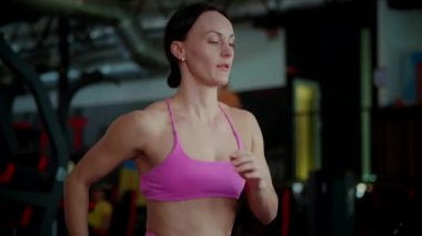 İyi aydınlatılmış bir spor salonundaki modern bir koşu bandında yoğun bir şekilde koşan pembe atletik giysili fit bir kadın. Odaklanmış kardiyo seansı: tanımlanmış sırt kasları olan atletik bir kadın kapalı alanda koşuyor. Parlak spor salonunun pencerelerinin altında adım adım ezici hedefler