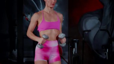 Kadının güzel bir atletik vücudu var ve dambılları güçlü bir şekilde kaldırıyor. Pembe bluzlu kadın antrenör ve şortlu spor salonunda dambıllarla çömelme hareketi yapıyor. Kadın sporcuların aktif fitness egzersizleri.
