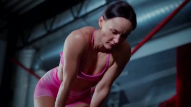 Pembe atletik giysili kadın, yoğun bir antrenmanın ardından oldukça yorgun görünüyor. Kendini işine adamış bir sporcunun loş bir spor salonunda soluklanmasına yakın çekim. Zinde bir kadının yüzünde azim ve kararlılık var. İyileşiyor.