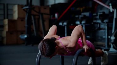 Kadın yoğun bir şekilde paralel şınav çekiyor loş bir spor salonunda vücudunun üst kısmına odaklanıyor. Paralel hareketler, sıkı bir antrenman sırasında oyulmuş sırt kasları. şınav formunun en zor kısmını ezip geçmek.