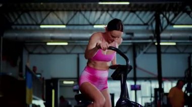 Pembe atletizm giymiş bir kadın loş bir spor salonunda hava bisikleti kullanıyor. Kararlı bir sporcunun durağan bir bisiklette ter içinde kalması. Formda bir kadının sıkı bisiklet sürerken tüm vücudunu çekmesi, fonksiyonel fitness ayarına odaklanmış..