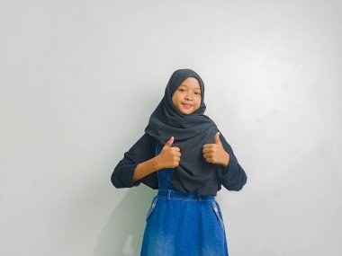 Siyah tesettürlü bir kız, iki başparmağı havada duruyor, beyaz bir arka planda izole edilmiş..