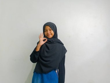 Siyah başörtüsü takan bir kız, bir eli kalçasında, diğeri ise beyaz arka planda izole bir şekilde 