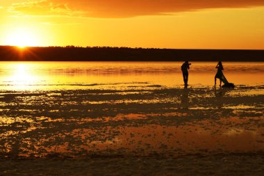 Bir fotoğrafçının siluetleri ve gölde elbise giymiş bir kız. Güneş ufukta batıyor ve gökyüzünün sudaki yansıması..