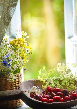 Bahçeye bakan eski bir ahşap pencerede olgun kırmızı kirazlar ve kır çiçekleri. Yaz havasında klasik bir fotoğraf. Yumuşak seçici odak.