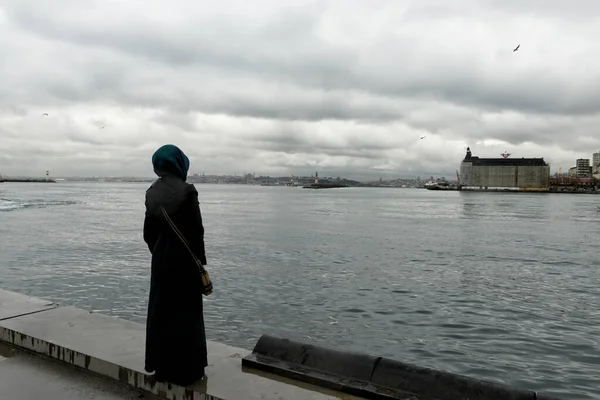 Geleneksel elbiseli Müslüman bir kadın, bulutlu ve dramatik bir havada Boğaz kıyısında duruyor..