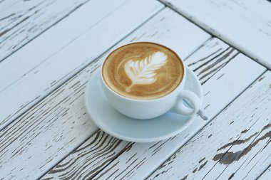 Beyaz ahşap masadaki köpüklü tüy desenli bir bardak kapuçino..