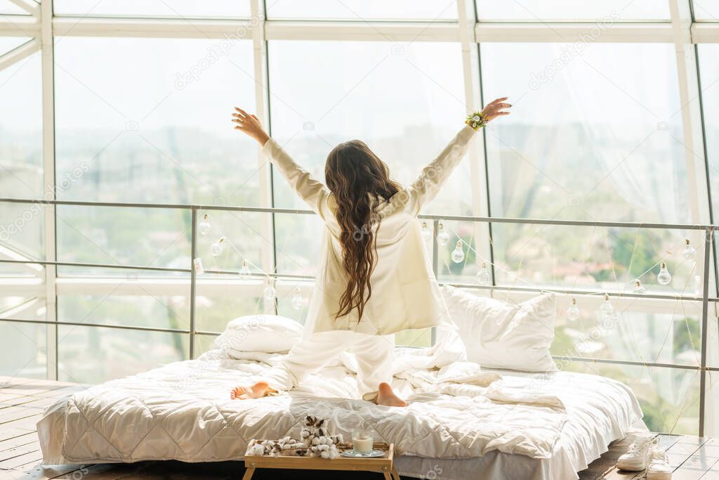 A cute young girl with long hair is lying on the bed overlooking a large bright window. The atmosphere of a cheerful bright morning.