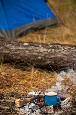 Sıcak içeceği olan mavi metal bardak ormandaki kamp ateşi közlerinde duruyor. Yangından yükselen duman, arka planda mavi çadır ve düşen kütük. Vahşi doğada rahat bir kamp ortamı.. 