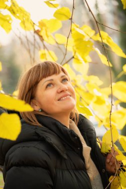 Kızıl saçlı bir kadın gülerek sonbahar havasının tadını çıkarıyor sarı ağaç yaprakları arasında parkta. Canlı sonbahar yeşillikleriyle çevrili yakın plan bir portre. 