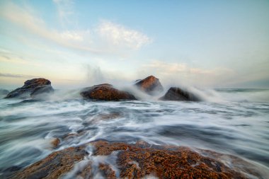 Uzun süreli deniz manzarası kıyı kayalarının etrafında dönen dalgaları yakalıyor. Suyun ve pastel gökyüzünün yumuşak hareketi doğa, seyahat ve rahatlama temaları için mükemmel sakin ve güçlü bir deniz manzarası yaratır..