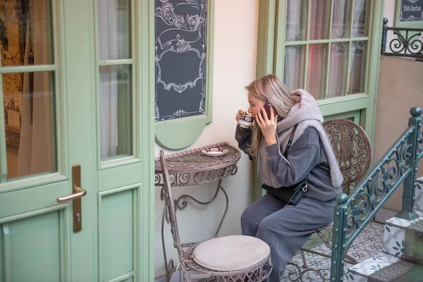 52 yaşında şık, sarışın bir kadın kahve içiyor ve zarif bir açık kahve masasında akıllı telefonuyla konuşuyor. Sıcak giyimli, rahat ve şehir atmosferinin tadını çıkarıyor, modern hayatları yakalıyor.