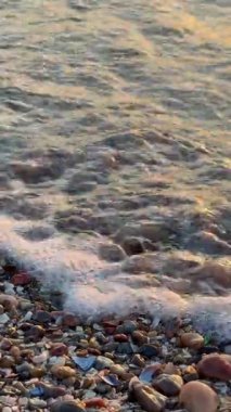 Altın saat boyunca plajda pürüzsüz çakıl taşlarının üzerinde köpük ve yansımalarla yıkanan küçük dalgaların yakın görüntüsü.