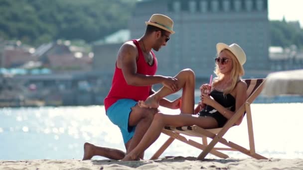 Man zastosowaniu ochrony przeciwsłonecznej na nogi dziewczyny na plaży — Wideo stockowe
