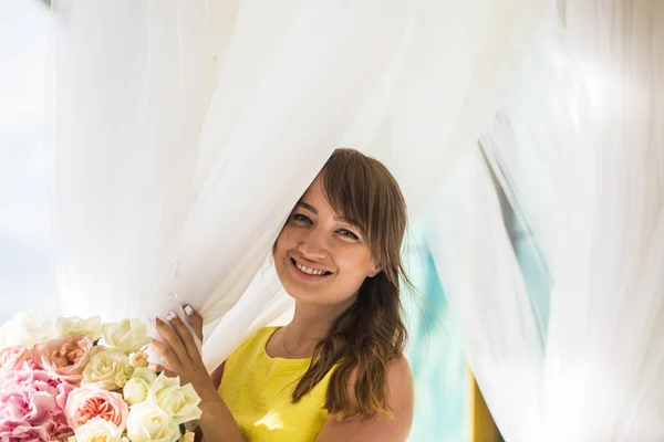 Vacker ung flicka i en gul klänning på terrassen med bakgrund av färska blommor — Stockfoto