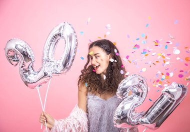 Beautiful young brunette girl with curly hair posing on a pink studio background with confetti and holding in her hand silver balloons from the numbers 2021, happy new year concept clipart