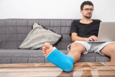 Evdeki kanepede dizüstü bilgisayarda çalışırken bileği burkulduğu için mavi bacağı kırık bir adam tedavi görüyor..