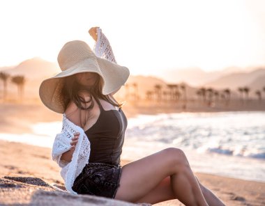 Güneşin batışında, kafasında şapka olan beyaz bir mayo giymiş güzel bir manken kumsalda poz veriyor.