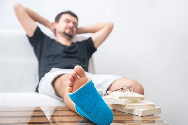 Man with broken leg in blue splint for treatment of injuries from ankle sprain reading books at the home rehabilitation.