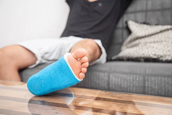 Man with broken leg in cast on couch at home. Sports injury concept.