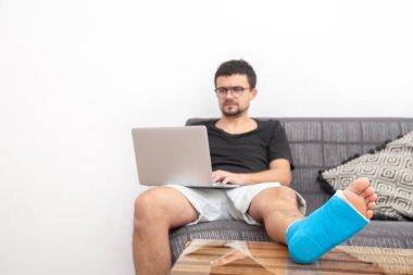 Evdeki kanepede dizüstü bilgisayarda çalışırken bileği burkulduğu için mavi bacağı kırık bir adam tedavi görüyor..