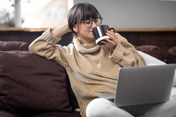 A young beautiful girl sits on the sofa at home with a cup of drink and cheerfully looks at the computer screen. The concept of video communication and communication at a distance.