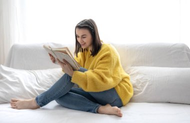 Sarı kazaklı şık kız evde kanepede otururken hevesle kitap okuyor..