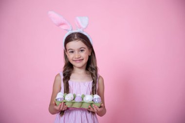 Paskalya tavşanı kulakları ve pembe stüdyo arka planında bir tepsi yumurtası olan neşeli küçük bir kız. Paskalya tatili konsepti.