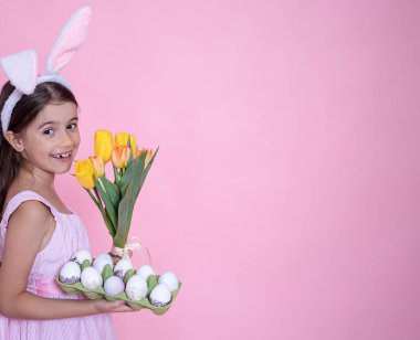 Paskalya tavşanı kulaklı küçük kız pembe stüdyo arka planında bir buket lale ve bir tepsi yumurta tutuyor..