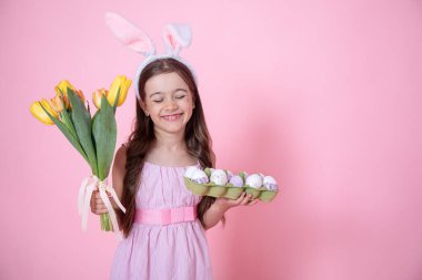 Paskalya tavşanı kulaklı neşeli küçük kız elinde bir buket lale ve pembe bir stüdyo arkasında bir tepsi yumurta tutuyor..