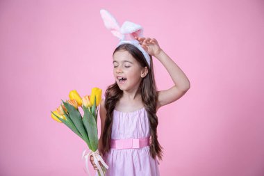 Paskalya tavşanı kulaklı neşeli küçük kız pembe stüdyo arka planında bir buket lale tutuyor..
