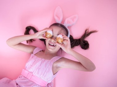 Paskalya tavşanı kulaklı küçük kız pembe stüdyo arka planında duran neşeli paskalya yumurtalarıyla poz veriyor..
