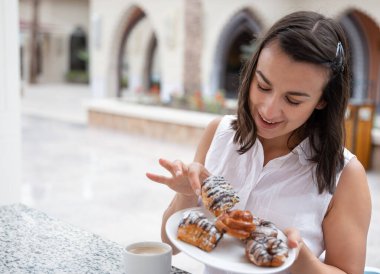 Dışarıdaki terasta çöreklerle sabah kahvesini yudumlayan neşeli genç bir kadın. Tatil ve eğlence konsepti.