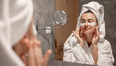 Kafasında havluyla bornoz olan bir kız banyoda aynanın önünde gözlerinin altına yamalar yapıştırıyor..