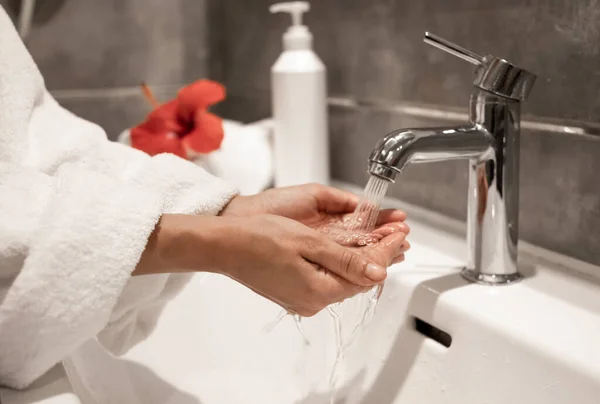 A woman in a robe washes her hands under running water from a tap.