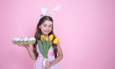 Paskalya tavşanı kulaklı küçük kız pembe stüdyo arka planında bir buket lale ve bir tepsi yumurta tutuyor..