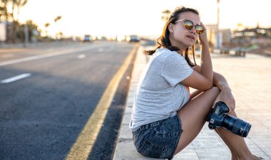 Yazın şortlu ve tişörtlü genç bir kadın gün batımında profesyonel bir kamerayla kaldırımda oturuyor..