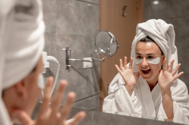 Kafasında havluyla bornoz olan bir kız banyoda aynanın önünde gözlerinin altına yamalar yapıştırıyor..