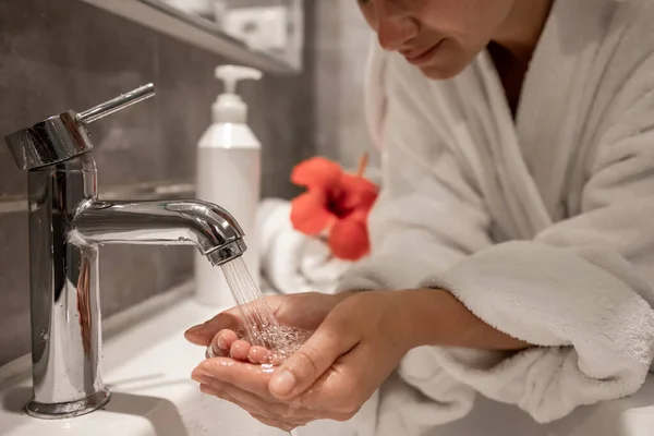 A woman in a robe washes her hands under running water from a tap.