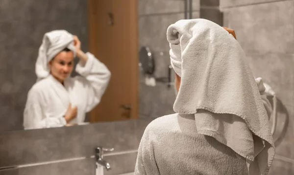 A young woman in a robe and with a towel on her head looks at herself in the mirror in the bathroom.