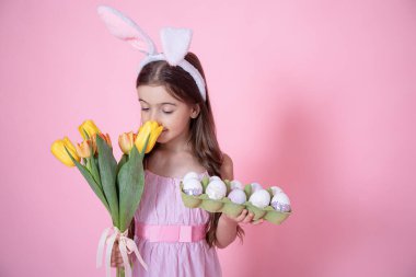 Paskalya tavşanı kulaklı küçük bir kız ve elinde bir tepsi yumurta pembe stüdyo arka planında bir buket lale kokluyor..