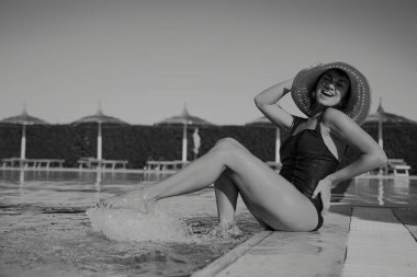 Şapkalı baştan çıkarıcı neşeli kız havuzun kenarında otururken ayaklarını ıslatıyor. Tatil ve eğlence konsepti. Siyah beyaz fotoğraf.