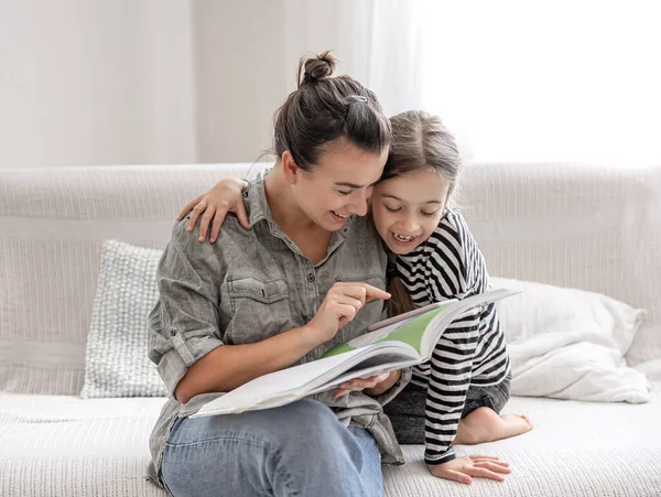 Cheerful mom and daughter are resting at home, reading a book together. The concept of a happy family and friendly relations.