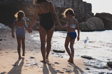 Anne ve iki küçük kız deniz kıyısında mayo giyip el ele tutuşarak yürüyorlar. Vee arkadan..